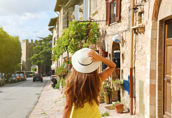 Beautiful young woman with yellow dress and hat walking in typical Assisi street, Italy. Rear view of happy cheerful girl visiting central Italy.