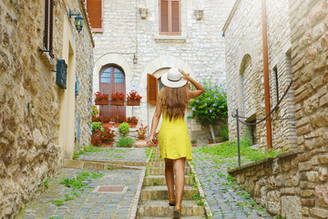 Curious young woman with yellow dress and hat goes upstairs in medieval street in Tuscany, Italy. Rear view of happy cheerful girl visiting central Italy.