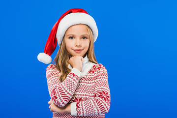 front view of smiling kid in sweater and santa hat isolated on blue