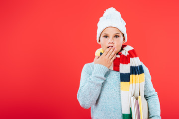 front view of surprised kid in hat and scarf covering mouth with hand isolated on red