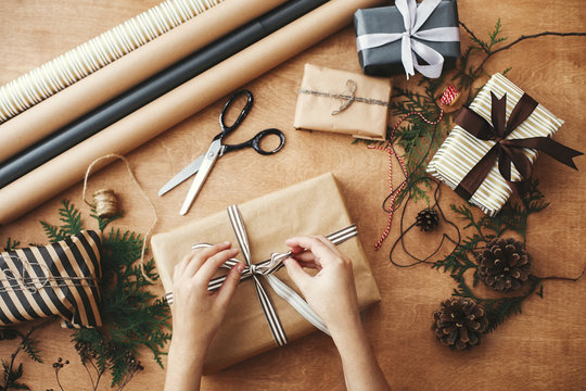 Merry Christmas, flat lay. Hands wrapping stylish christmas gift box in craft paper and scissors, rustic  presents, thread, pine branches and cones on wooden table.