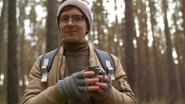 Portrait of young Caucasian hiker holding metal thermo cup and looking at camera while standing in forest on autumn day