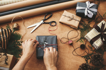 Wrapping christmas gifts flat lay. Hands wrapping stylish christmas gift box in black paper and scissors, rustic presents, thread, pine branches and cones on wooden table - obrazy, fototapety, plakaty