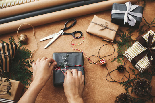 Wrapping christmas gifts flat lay. Hands wrapping stylish christmas gift box in black paper and scissors, rustic presents, thread, pine branches and cones on wooden table