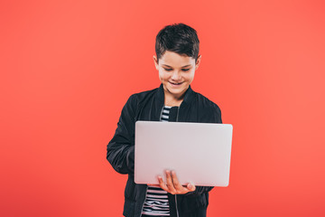 front view of smiling kid in jacket using laptop isolated on red