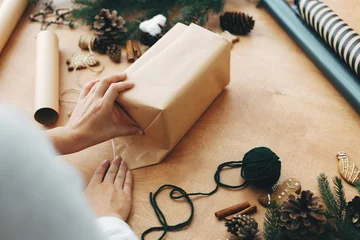 Fotobehang Hands wrapping christmas gift in craft paper and pine branches, cones, gingerbread cookies, thread, cinnamon on rural wooden table. Wrapping stylish christmas gifts concept © sonyachny