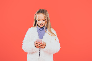 front view of smiling kid in autumn outfit using smartphone isolated on red