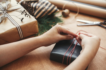 Hands wrapping stylish christmas gift box in black paper and scissors, rustic presents, thread, pine branches and cones on wooden table. Wrapping christmas gifts