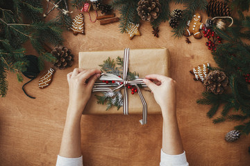 Merry Christmas flat lay. Hands holding stylish rustic christmas gift and pine branches, cones,...