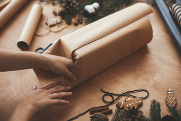 Hands wrapping christmas gift in craft paper and pine branches, cones, gingerbread cookies, thread,...