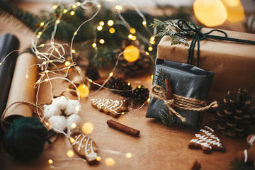Stylish rustic christmas gift in black wrapping paper with cinnamon decoration on background of golden lights, pine branches, cones, gingerbread cookies and cotton. Merry Christmas