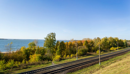 Fototapeta na wymiar Railway on the background of the river Bank on an autumn Sunny day. Beautiful yellow-blue panorama of trees by the water.