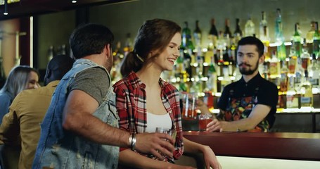 Beautiful young Caucasian woman standing at the bartender and drinking cocktail, then handsime man coming to her as wanted to acquaint with her, they talking, smiling and drinking.