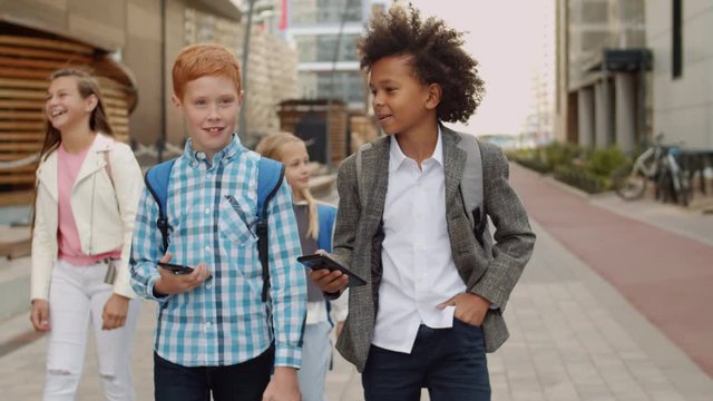 Medium shot of five diverse classmates with backpacks going out of school and communicating. Two boys using phones on foreground