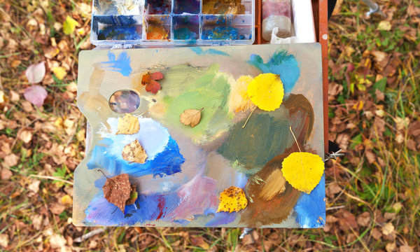 Artistic wooden palette with autumn colors paints and yellow leaves on background of fallen foliage. Autumn oil painting in open air