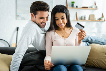 african american woman holding credit card and looking at laptop with man