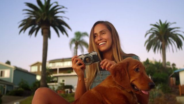 Young hip girl shooting photos with film camera and golden retriever puppy at sunset