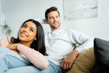 smiling african american woman lying on legs of handsome man in apartment