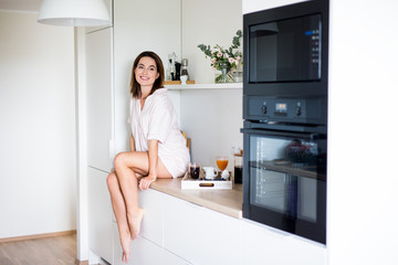 breakfast concept - woman in pajamas sitting in modern kitchen