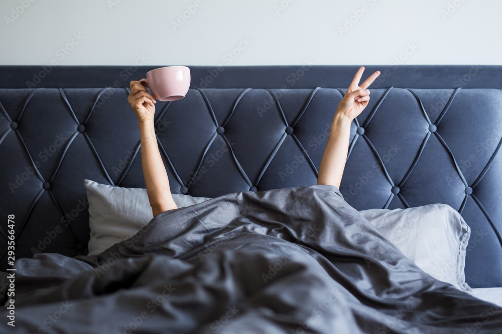 Wall mural good morning concept - female hands with coffee mug and victory sign sticking out from the blanket