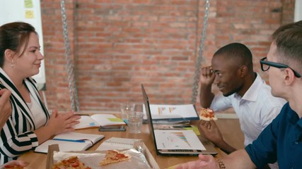 Diverse Group of People Eats Pizza in Loft Office. Business Team Enjoy Eating Lunch Together at Workplace with Laptops. Food Delivery Service and Catering at Work. 4K Gimbal Pull out Medium Shot
