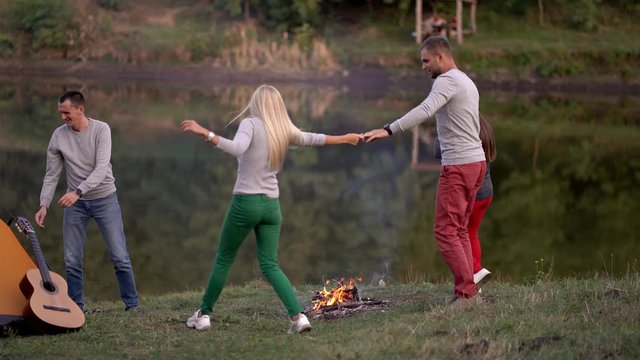 Four friends are running around the fire. A round dance is carried around a bonfire on the river bank. Picnic in nature, fun. Casual clothes, summer time, sunset light. Be happy, young and free.