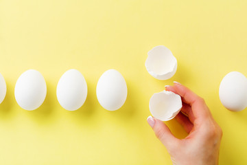 White raw chicken eggs lying on yellow light background and female hand holds broken egg. Top view.