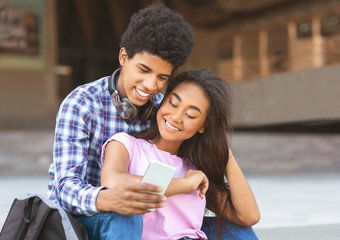 Smiling students scrolling photos in smartphone together sitting outdoors