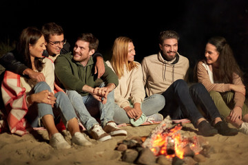 leisure and people concept - group of smiling friends sitting at camp fire on beach at night
