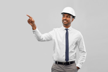 architecture, construction business and people concept - smiling indian male architect in helmet pointing finger to something over grey background