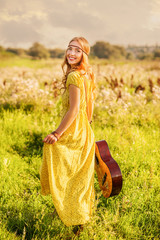 Happy girl in bohemian style yellow dress with guitar on the field turned back at sunset