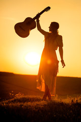 siluette of woman wearing a bohemian style holding a guitar on a field at warm light of sunset