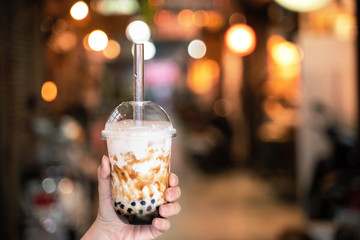 Young woman is holding, drinking brown sugar flavored tapioca pearl bubble milk tea with glass straw in night market of Taiwan, close up, bokeh