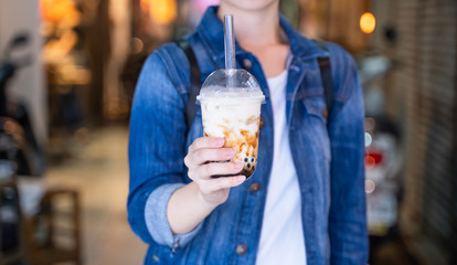Young woman is holding, drinking brown sugar flavored tapioca pearl bubble milk tea with glass straw in night market of Taiwan, close up, bokeh