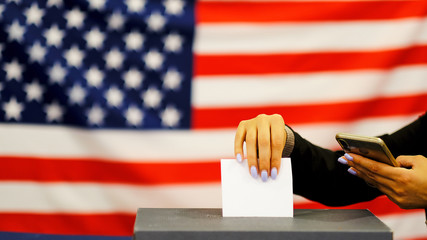 woman putting a ballot in a ballot box on election day. Close up of hand with white votes paper on...