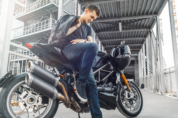 Fototapeta na wymiar Handsome biker sitting on black motorcycle on urban background at parking.