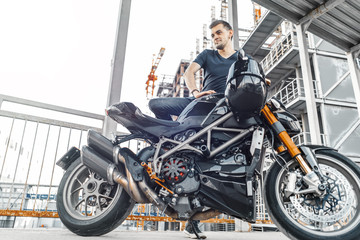 Fototapeta na wymiar Handsome biker sitting down on black motorcycle on urban background at parking.