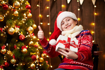 Cheerful santa helper girl with gift box sitting indoor near decorated xmas tree with lights, dressed in red sweater - Merry Christmas and Happy Holidays!