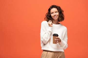 smiling shy woman holding coffee to go isolated on orange