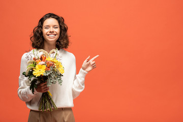 happy woman in autumnal outfit holding bouquet of flowers and pointing with finger isolated on orange