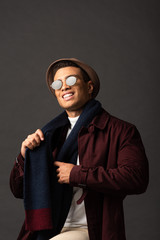 smiling elegant mixed race man in scarf, sunglasses and hat on black background
