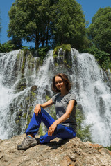 Young traveling woman near waterfall. Calm female tourist sitting on stone near waterfall and looking at camera