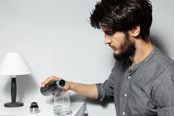 Portrait of young bearded guy with disheveled hair, pours water in glass from steel thermo bottle, on background of grey.