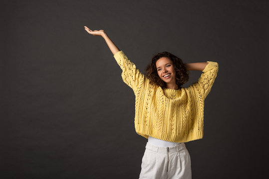 Smiling Curly Woman In Yellow Knitted Sweater With Hands In Air On Black Background