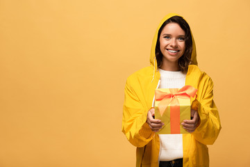 happy curly woman in yellow raincoat holding gift box isolated on yellow