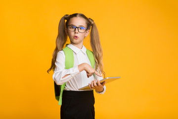 Confused little schoolgirl doing homework on digital tablet, yellow background