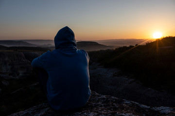 The guy is watching the dawn in the highlands. A magnificent landscape with a man in a hood. The athlete got up in the morning for training and looks at the mountain from the cliff.