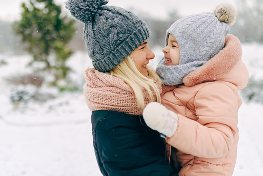 Outdoor portrait of beautiful happy woman holding her toddler girl, smiling and laughing together during walking in the park on snowing day. Christmas mood. Family portrait. Motherhood and childhood