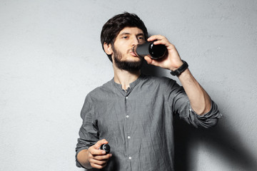 Portrait of young bearded guy, drinking water from black stainless bottle, on textured background of grey.