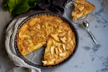 Apple pie in a round shape, decorated with fan-laced apple slices from the top, next to a cup of hot tea, autumn leaves, all on a light, marbled gray background. 